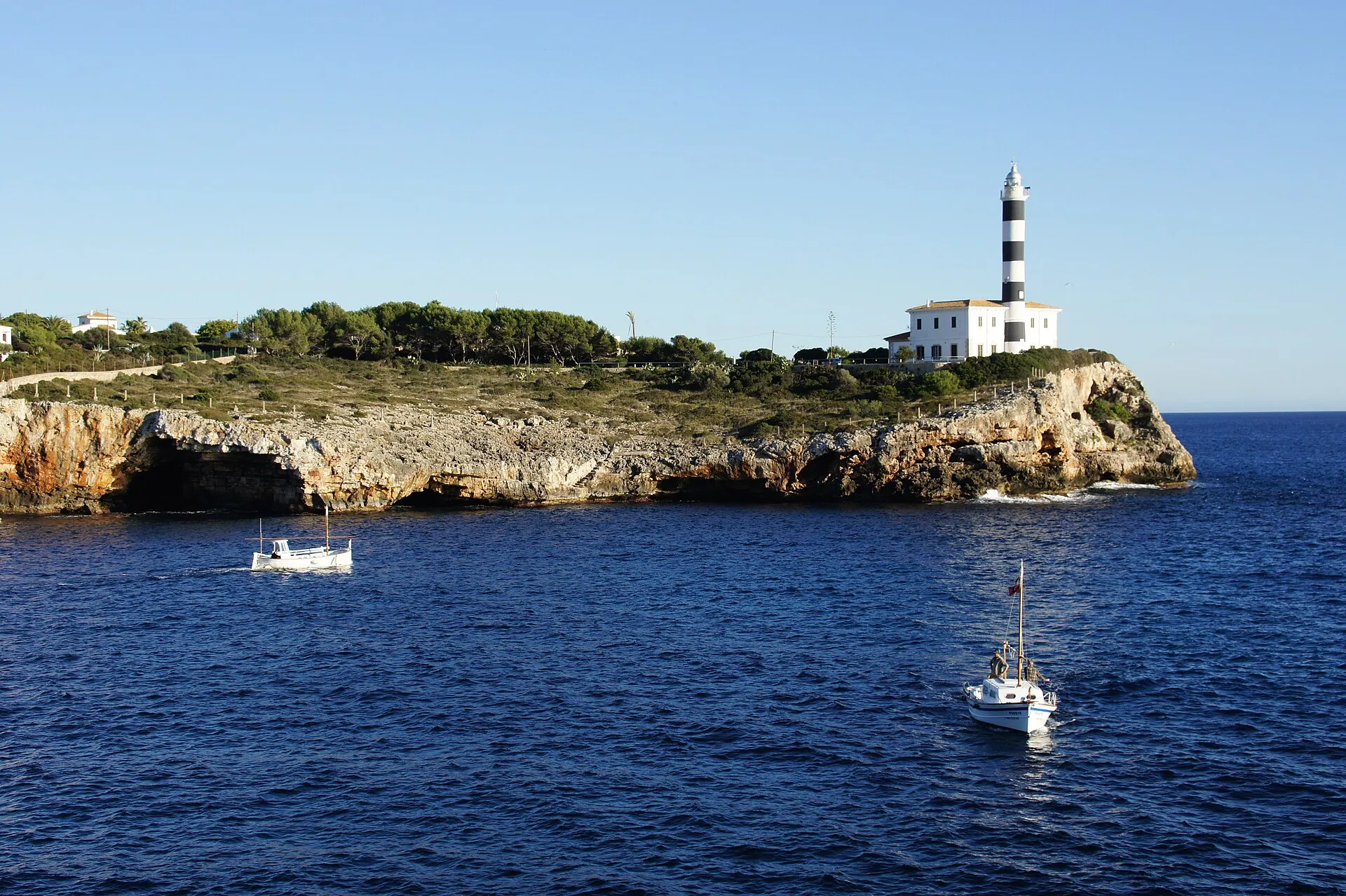 The banded lighthouse at the mouth of Portocolom harbour, boats passing on a clear evening.